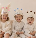 coroa-personalizada-nome-algodao - PIPÁPÁ - Amor em Madeira Three smiling toddlers in cream outfits wear paper crown hats with pom-poms, labeled Laura, Rodrigo, and Félix.