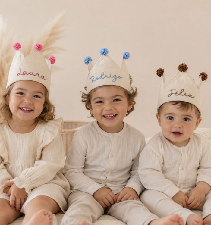 Three smiling toddlers in cream outfits wear paper crown hats with pom-poms, labeled Laura, Rodrigo, and Félix.