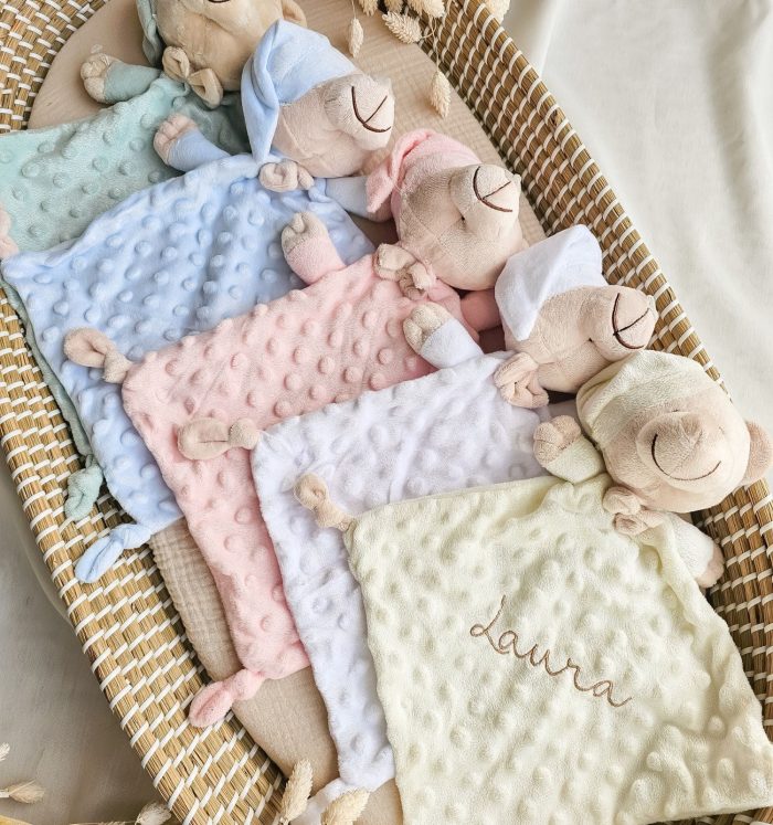 Soft pastel baby blankets with fuzzy plush sheep tucked in a wicker bassinet; colors blue, pink, white, and cream create a cozy nursery scene.
