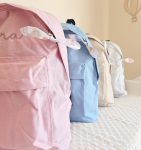 31 - PIPÁPÁ - Amor em Madeira Row of pastel backpacks in pink, blue, and beige lined up on a white quilted bedspread.