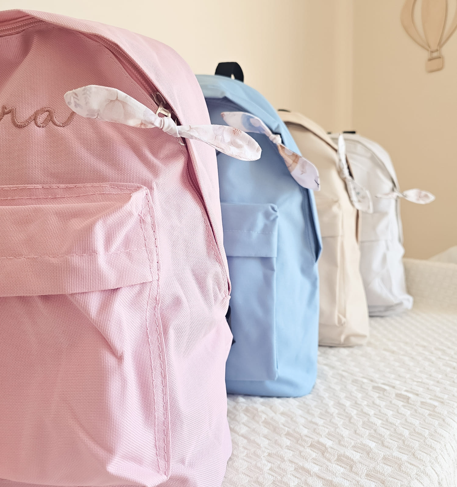 31 - PIPÁPÁ - Amor em Madeira Row of pastel backpacks in pink, blue, and beige lined up on a white quilted bedspread.