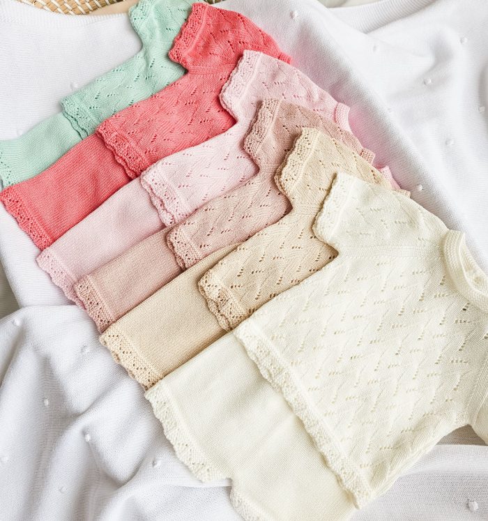 Stack of pastel knit baby sweaters with lace edges on a white textured blanket surface.