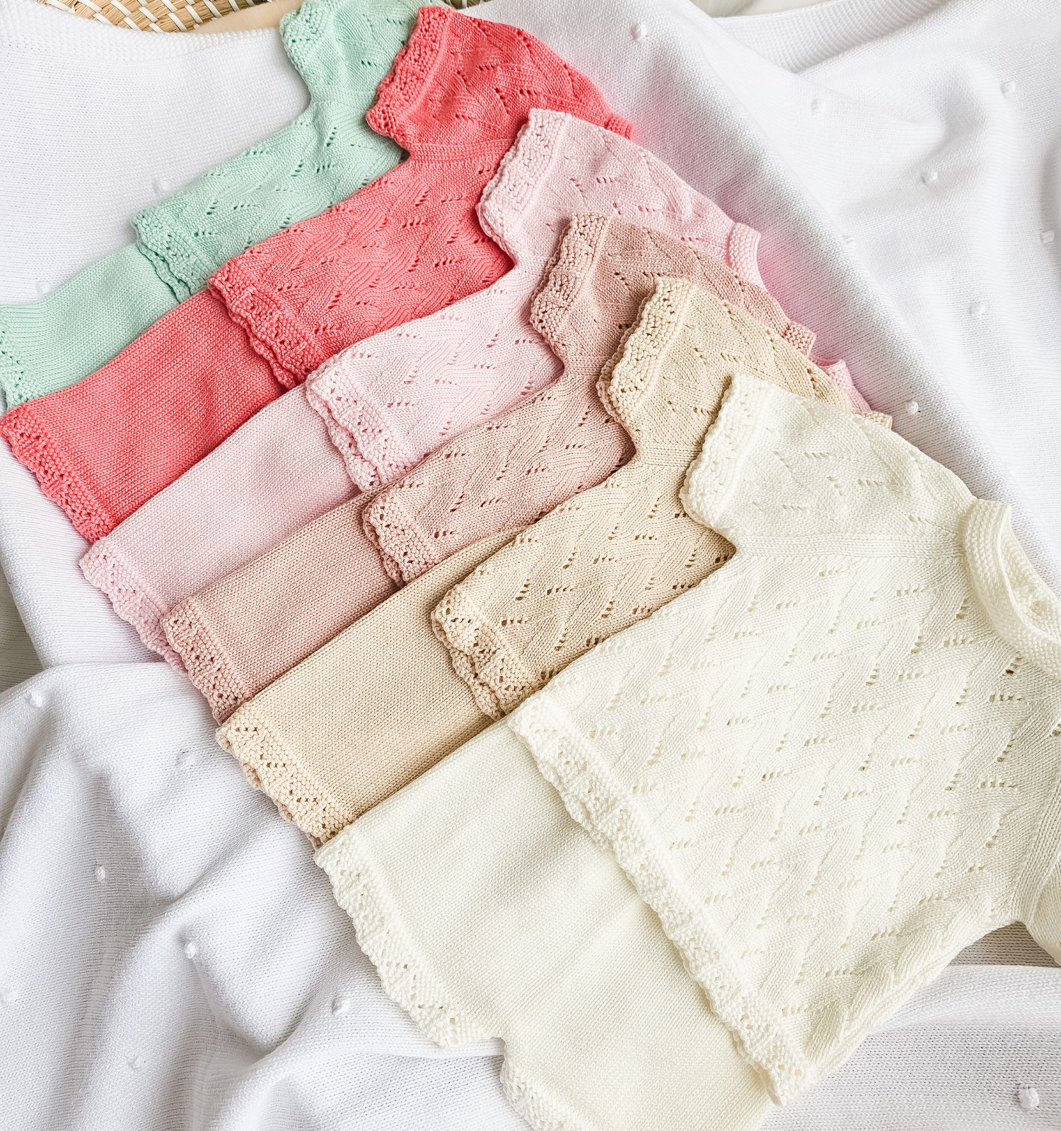 conjunto-malha-verao-bebe - PIPÁPÁ - Amor em Madeira Stack of pastel knit baby sweaters with lace edges on a white textured blanket surface.