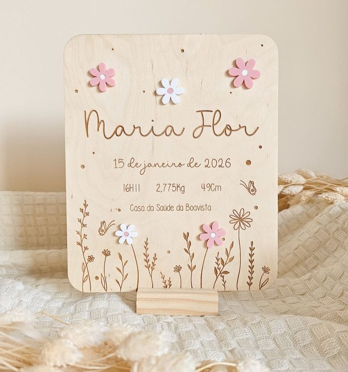 Wooden birth announcement board with engraved name 'Maria Flor', date, measurements, and floral decorations; pink and white flower pins on top.